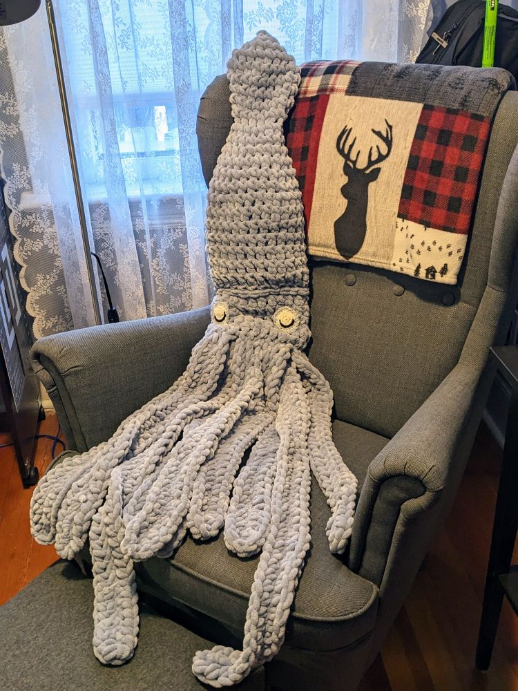 A light blue-grey, crocheted giant squid reclines on a dark grey armchair.  There is also a throw blanket with red buffalo plaid and deer silhouettes on the top of the arm chair.