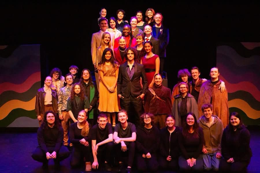 The cast and crew of Romeo & Juliet are arranged in an inverted T shape. Romeo & Juliet are surrounded by the Prince and the Montague & Capulet families, and Count Paris. To the left are Benvolio, Mercutio, Balthazar, & the Friar Lawrence. To the right are Tybalt & other members of House Capulet. The costumes are modern, but timeless in rich shades of red, gold, royal blue, & emerald. The crew kneels at ground level, clad all in black. 