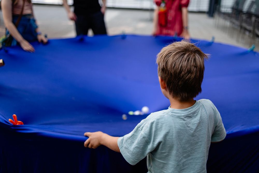 A round blue cloth with little white balls rolling towards its depressed center. An audience gathering around the installation.