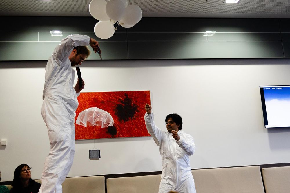 A person standing on a table, reaching up to the balloon with a pair of scissors and cutting the string between balloon and the attached model.