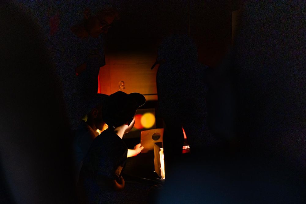 A picture that is mostly dark. Near the image center, two light blobs in red and yellow color as projected into a wall and appear distinctively off-set. Two kids' heads are barely visible in the dark, looking into the direction of the projection.