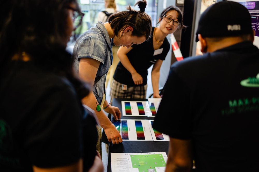 A teen bending over print-outs of rainbow-colored stripes: a star's spectrum. More people crowd around the same table, the picture is taken peeking through a small gap between two of them.
