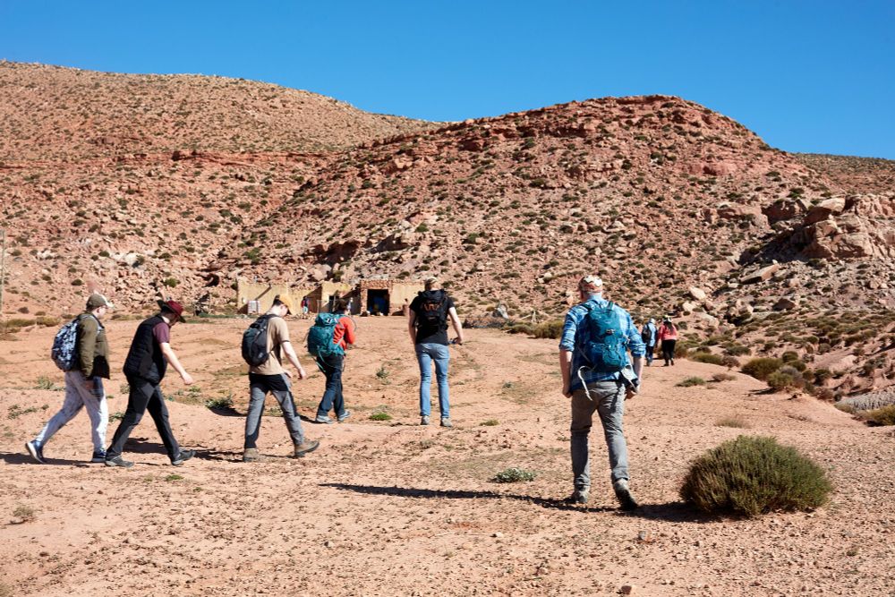 A group of people in hot weather gear and backpacks walk towards an arid hillside covered in rocks and scrub. A flat building sits at the base of the hill.