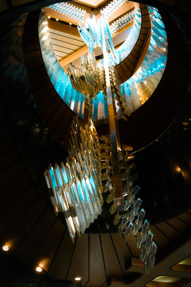 A photo of the several meters long chandelier in The New Stage Theatre in Prague. It’s made of two metal rods coming from the ceiling with different clear glass elements attached to it which look like shards. They are at the bottom, the middle and the head part of the rods. The chandelier looks a little like a surreal snow flake or like a frozen art piece
