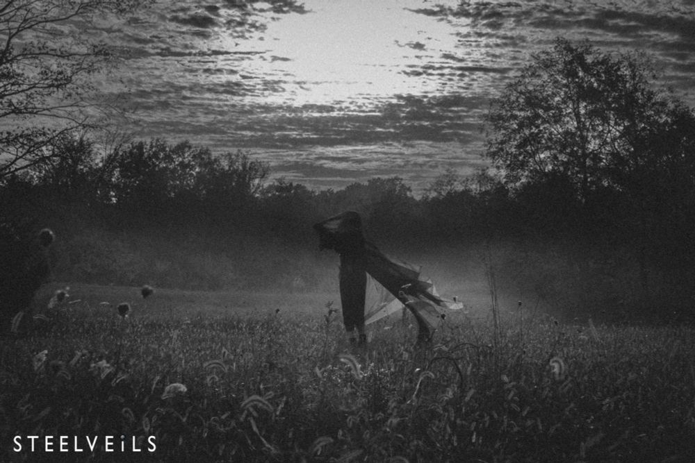 Black and white image of a figure draped in cloth moving across a dusty field with trees in the distance, and good cloud detail in the sky. 