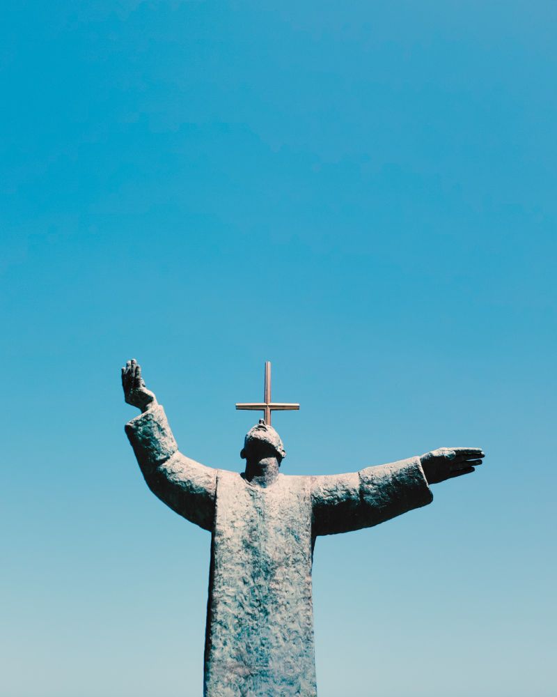 Statue of a Spanish priest with a cross behind him in a blue sky.
