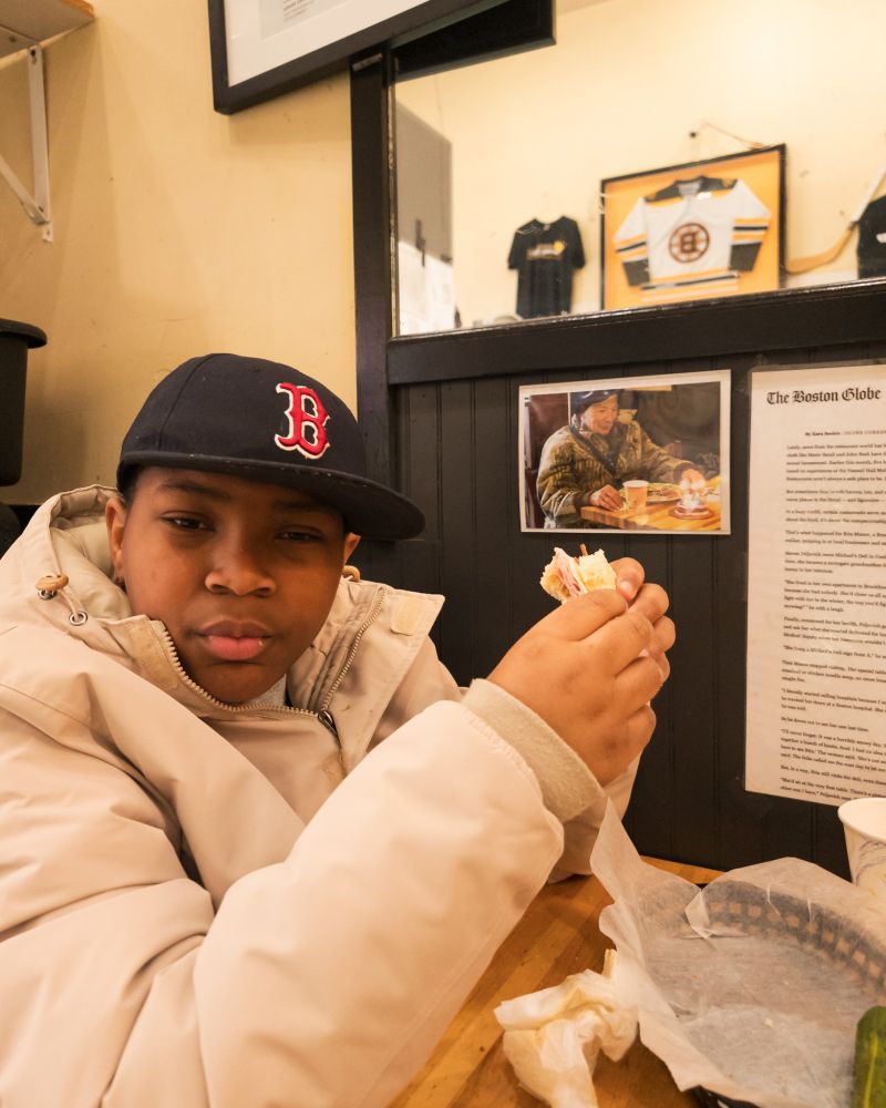 A little boy in a Red Sox hat eating a sandwich.
