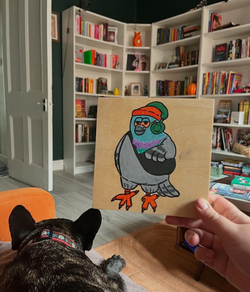 A wooden canvas with a cartoon pigeon on it photographed next to a sleeping frenchie.