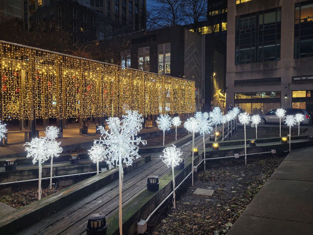 X-shaped wooden bridge lined with short poles topped with lighted white snowflake globes, with a curtain of lights in the background