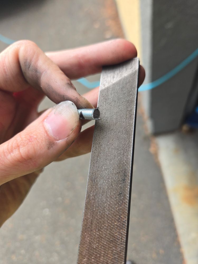 A picture of a no-longer-too-long machine screw, with rough edges, being smoothed out on a file. Your goal isn't to take away too much material - just smooth off the roughness and try to avoid damaging or altering the threads at all.