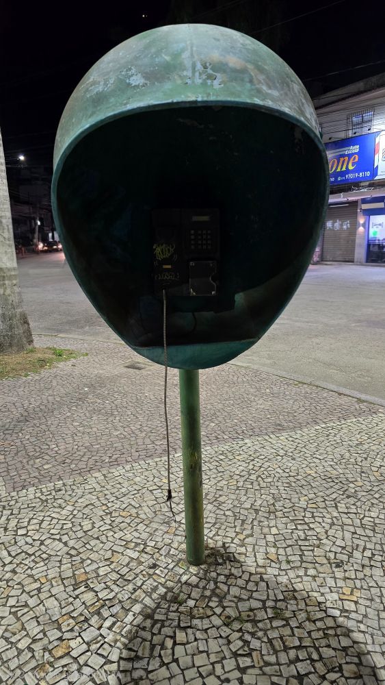 Vista frontal de um orelhão depredado, mas ainda com o telefone no lugar, embora o fone tenha sido arrancado. Em uma praça pública do Rio de Janeiro. 