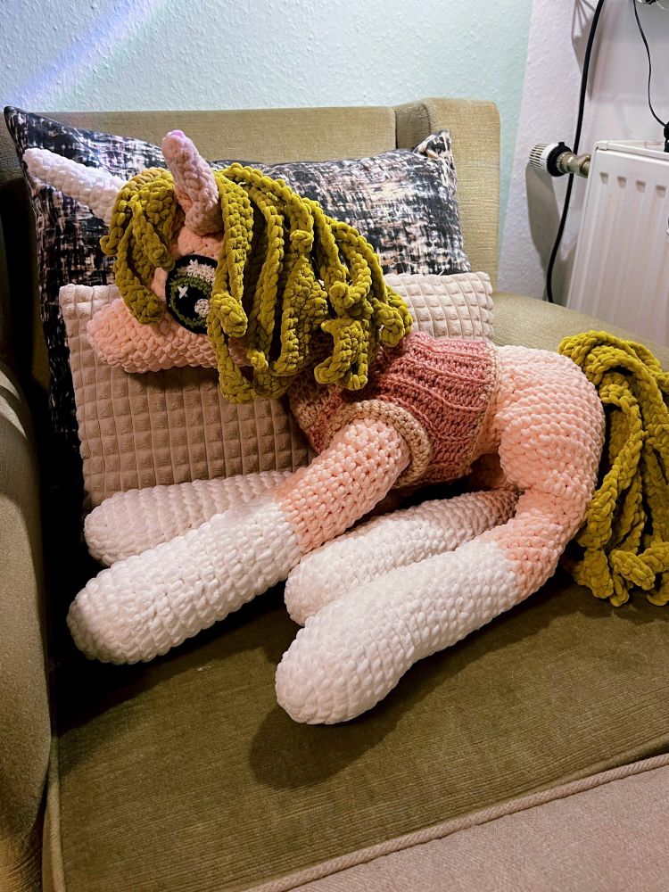 A large handmade crochet pony with a unicorn horn lies stretched out on a sofa, leaning on beige pillows. The pony is pink and white with green yarn hair and big green eyes.