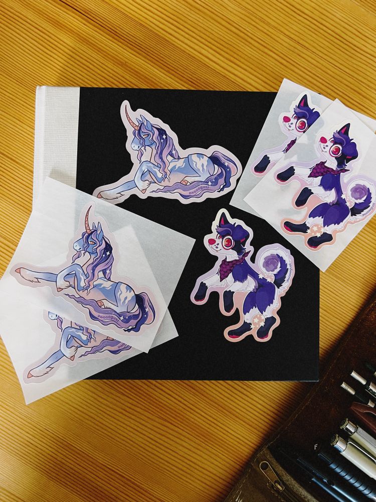 Photos showing a small giveaway set for Eurofurence: two colorful postcards, two matching stickers, and a standee with a character peeking over the top that displays QR codes for social media. The items are laid out clearly to show the artwork and quality.