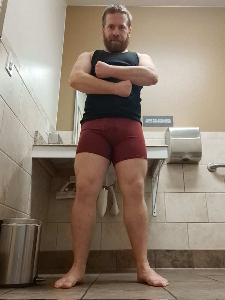 Photo of me in red boxer briefs and a black undershirt, in a gym bathroom. I am showing off my muscular arms and legs.