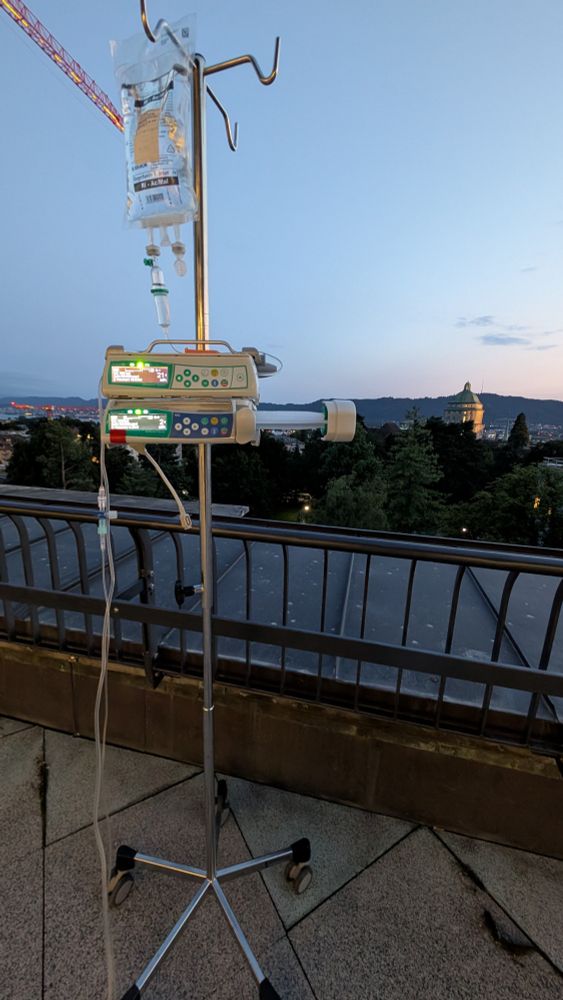 Ein Infusionsständer mit Infusionsbeuteln und einem Infusionspumpen-Gerät steht auf einem Balkon. Im Hintergrund ist eine Stadtlandschaft bei Sonnenuntergang zu sehen. Die Dächer der Gebäude und die Baumkronen sind von der Abenddämmerung umhüllt. Ein historisches Gebäude mit einer Kuppel ist in der Ferne erkennbar. Der Himmel hat einen sanften Farbverlauf von Blau zu Rosa und Orange.
