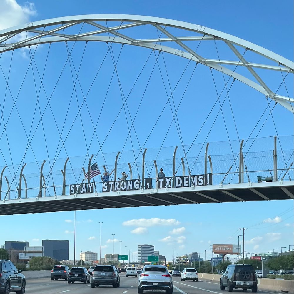People on a bridge over a freeway holding signs that read- “STAY STRONG, TX DEMS!” 