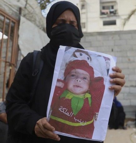 Zainab's mother is dressed in a black hijab and mask and holds a printed photo of a Zainab lying on her back with her hands up by her face, dressed in a red and green outfit, resembling a strawberry with a collar of green leaves. Zainab has chubby cheeks and brown eyes. 