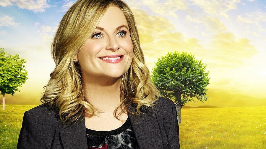 A white woman with blonde hair wearing a suit jacket stands smiling in front of a sunny yellow field with green trees.