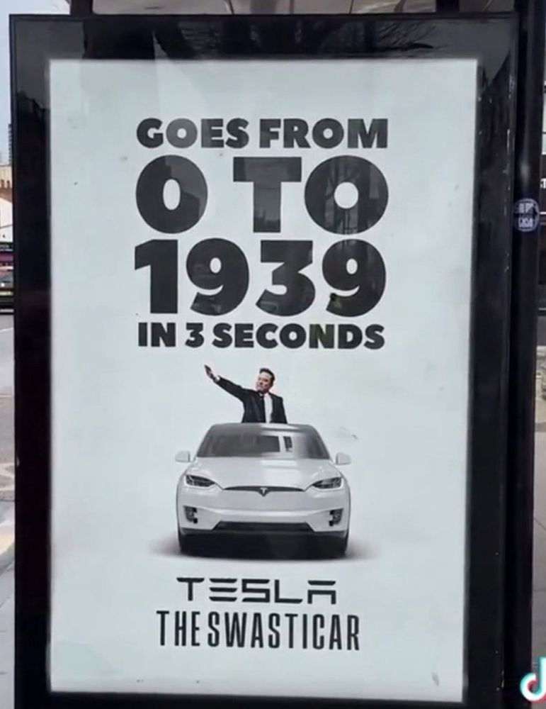 Sign saying :Goes from 0 to 1939 in 3 seconds - Tesla the Swasticar. Image of Elon Musk in a Tesla giving Hitler salute. 