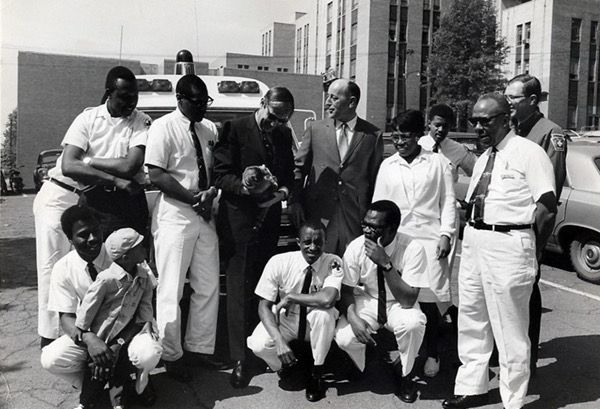 A group of black emergency medical technicians from Freedom House are gathered for a group shot in front of an ambulance. Almost all wear white suits. 