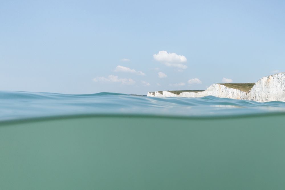 A photograph taken in the sea showing the white chalk cliffs at the Seven Sisters in East Sussex, an undulating blue sea under a clear sky, and that particular chalky green colour of the water beneath the surface. All the colours of summer in the sea. 