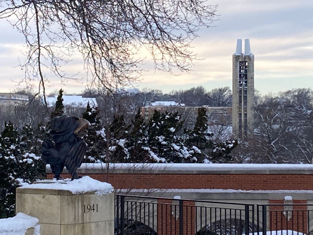 A picture of a statue of the 1941 Jayhawk overlooking a black fence in front of a red brick walkway. There are snowy trees, and the Jayhawk is facing the Campanile Bell Tower.