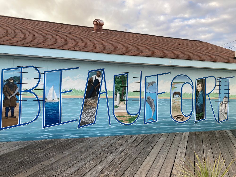 Picture of a mural reading “Beaufort” (pronounced Bewfurt) with a background consisting of the ocean and a low-laying island. Each letter contains a picture related to the town’s history. B has Blackbeard, E has a sailboat, A has a black man harvesting cotton, U has a white fence with the gate open, F has a dolphin, O has the Shackleford Banks horses and a crab, R has Rachel Carson wearing binoculars around her neck and leaning against a tree, and T has an old white house.