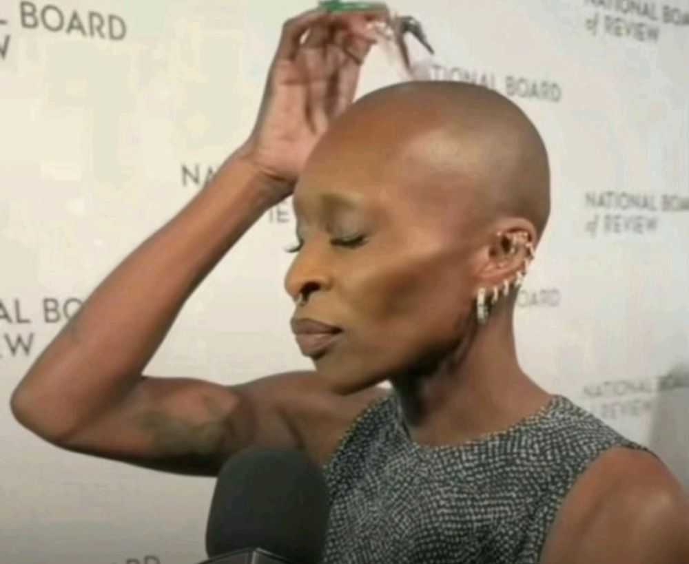 Image of Cynthia Erivo at a red carpet event, typing on the temple of her head with her nails like she's mentally calculating something. 
