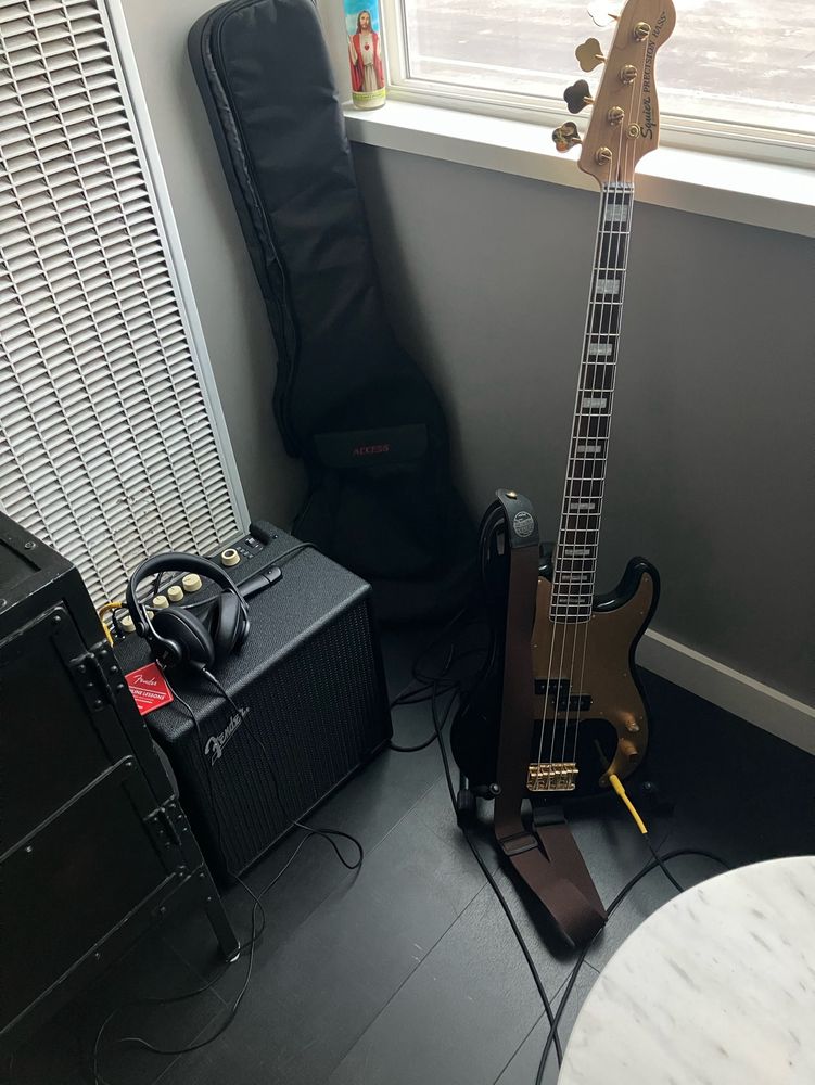 Squier electric bass next to amp with headphones in corner of apartment