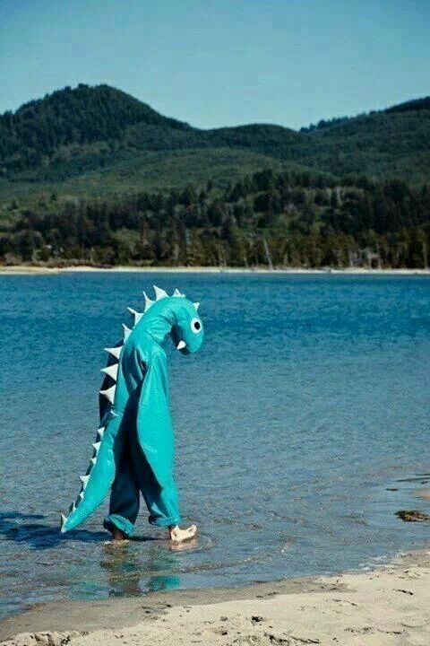 Pessoa com fato de dinossauro na água, com uma floresta atrás 