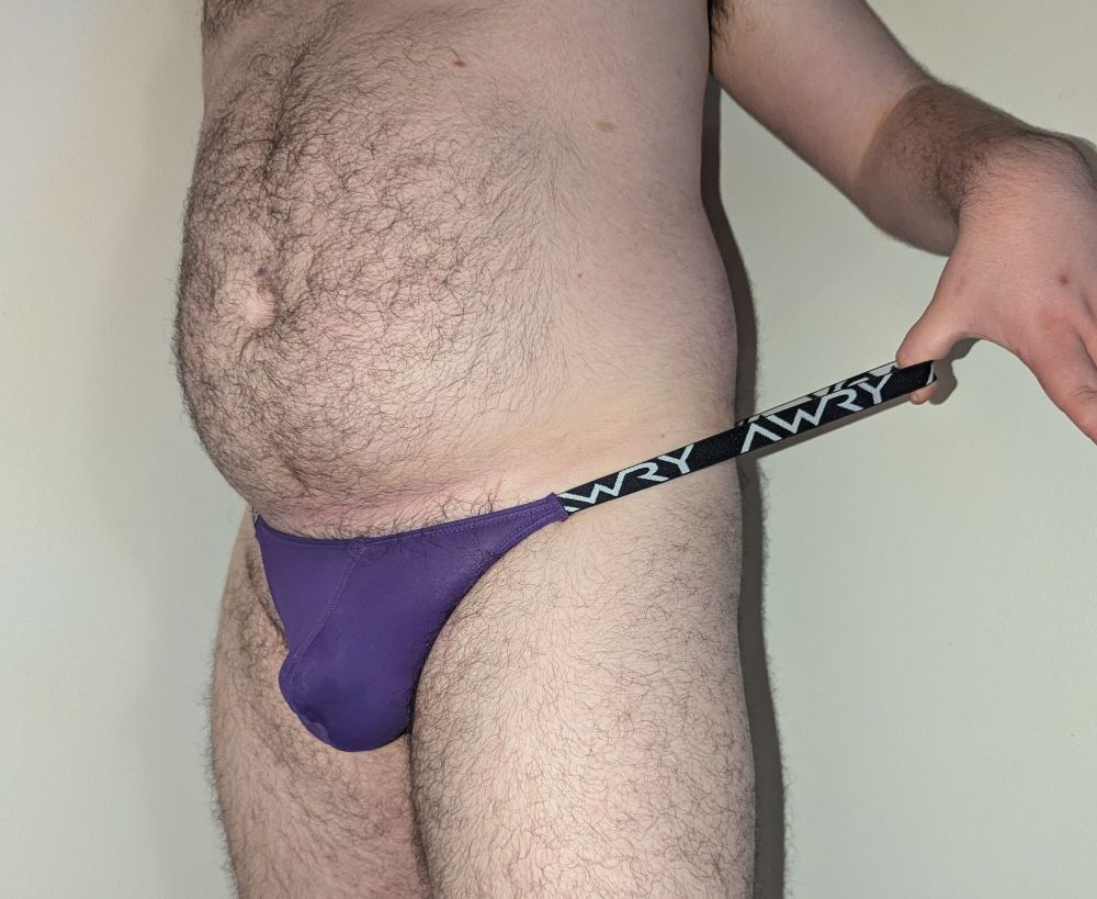 My lower torso, clad in only a purple thong. The logotype "Awry" is printed on the black and white waistband, which I'm pulling away from my waist with my outstretched hand.