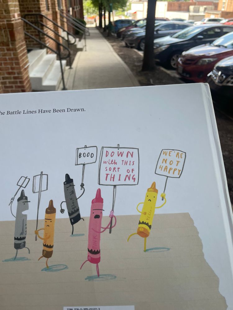The back cover of a hardcover copy of the children’s book ‘The Day the Crayons Quit’ with an illustration of anthropomorphic crayons holding protest signs like “we are not happy” and “down with this sort of thing.”