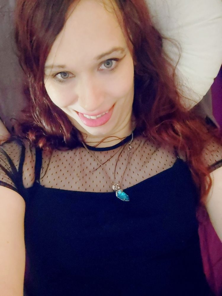 Selfie of Elaine in a smart gothic top and blue pendant, she's biting her tongue towards the camera with a mischievous grin.