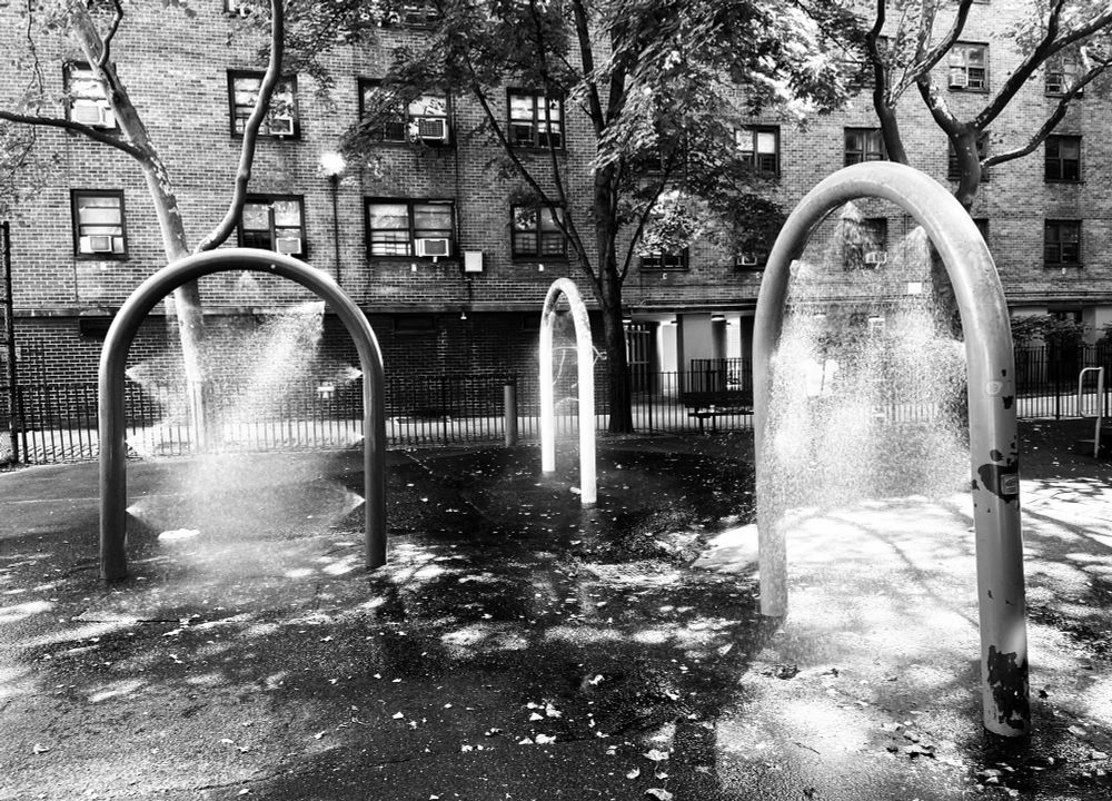 Black and white medium shot of a playground in between apartment buildings in Manhattan. There are three arches that emit water for cooling on hot days of summer.