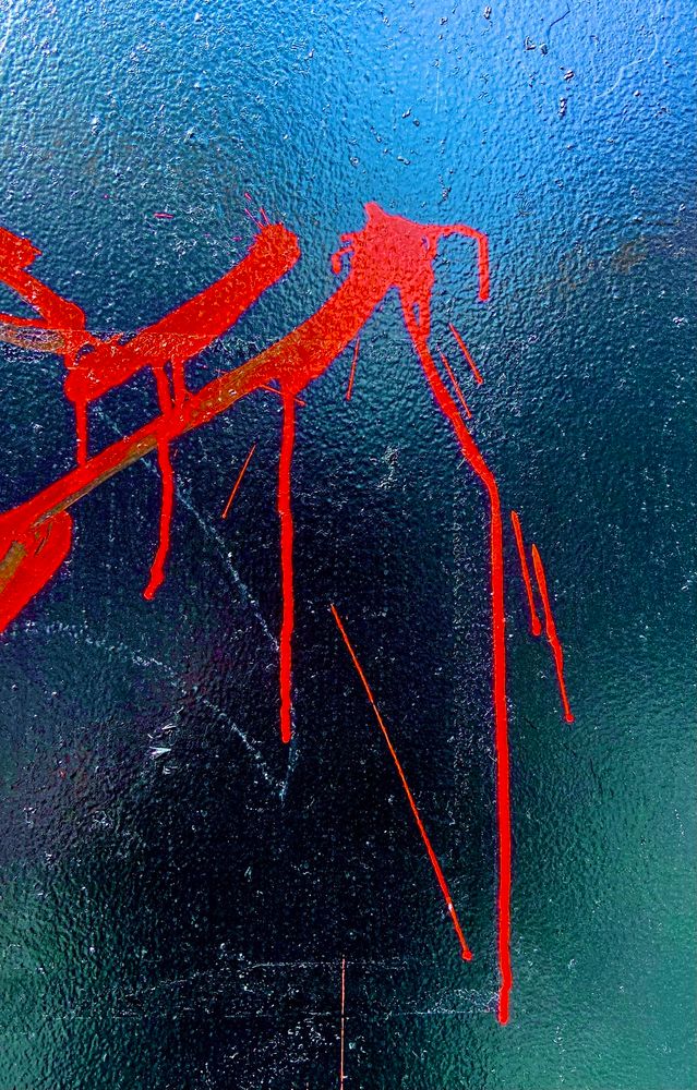 Detail of red graffiti lines on a wall painted in reflective black. In the light of the reflection, the vague visage of me taking the photo is visible.
