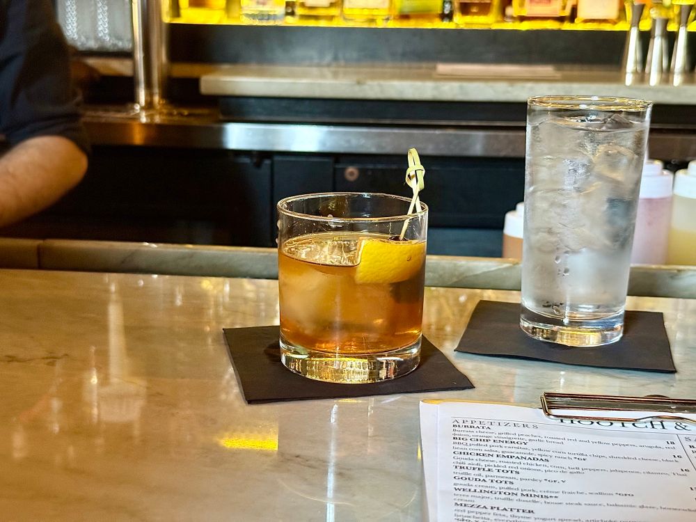 A cocktail garnished with a lemon peel sits on a black napkin atop a marble bar, next to a tall glass of ice water. The background shows a dimly lit bar with liquor bottles and a partial view of a bartender’s arm, while a printed appetizer menu is partially visible in the foreground.