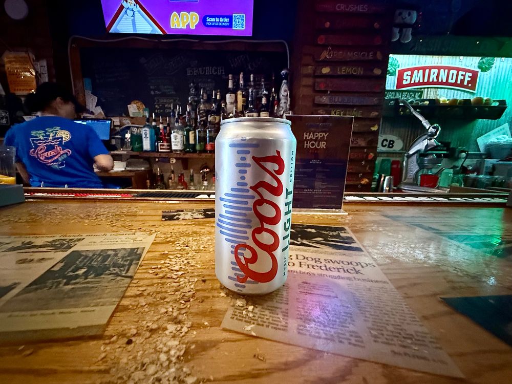 A Coors Light beer can with a red, white, and blue design stands on a wooden bar counter covered in old newspaper clippings and wood shavings. The background shows a colorful bar scene with liquor bottles, a bartender in a blue t-shirt, and signage for Smirnoff and Happy Hour.