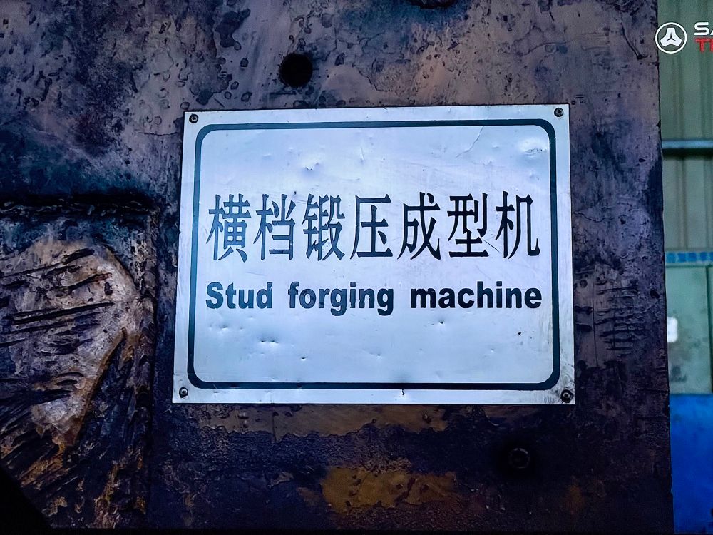 Close-up of a metal sign bolted onto an industrial machine, with Chinese characters and the English label “Stud forging machine” printed below. The surface around the sign appears rusted and worn, indicating heavy use in a manufacturing or metalworking environment.