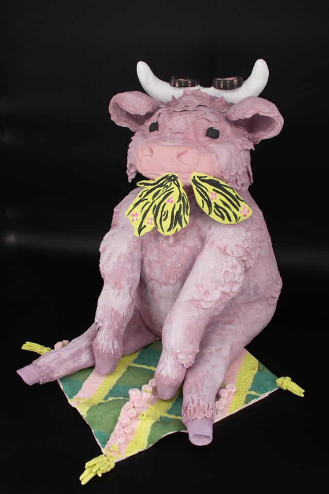 A large purple highland cow sculpture, sitting like a dog on a plaid rug that is meadow-like and munching on some chartreuse greens with pink flowers.