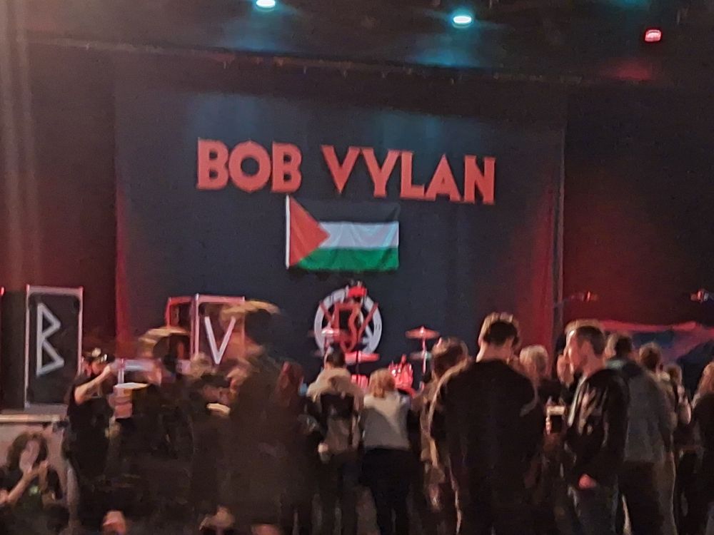 A gig venue starting to fill up. There is a Bob Vylan backdrop onstage with a Palestinian flag.
