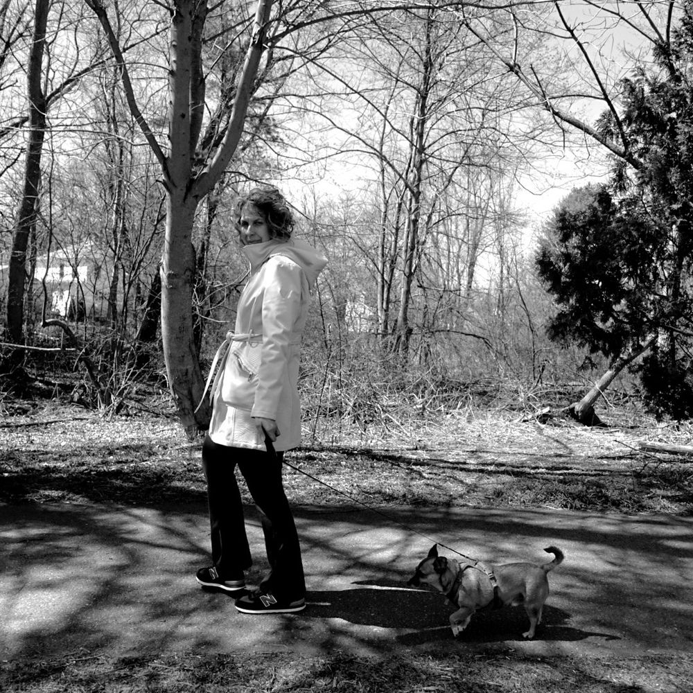 Woman walking small dog on shadow path wearing a white coat. 