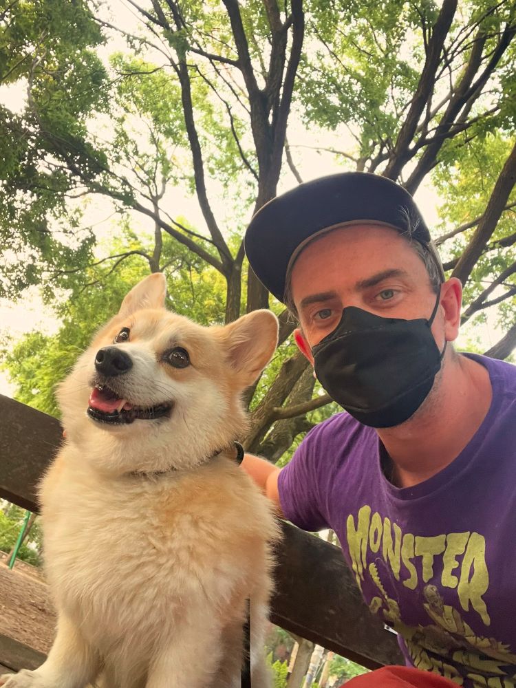 A cute corgi and his human dad pose on a bench. Corgdad is wearing a mask to protect himself from the smoky air caused by the wildfires.