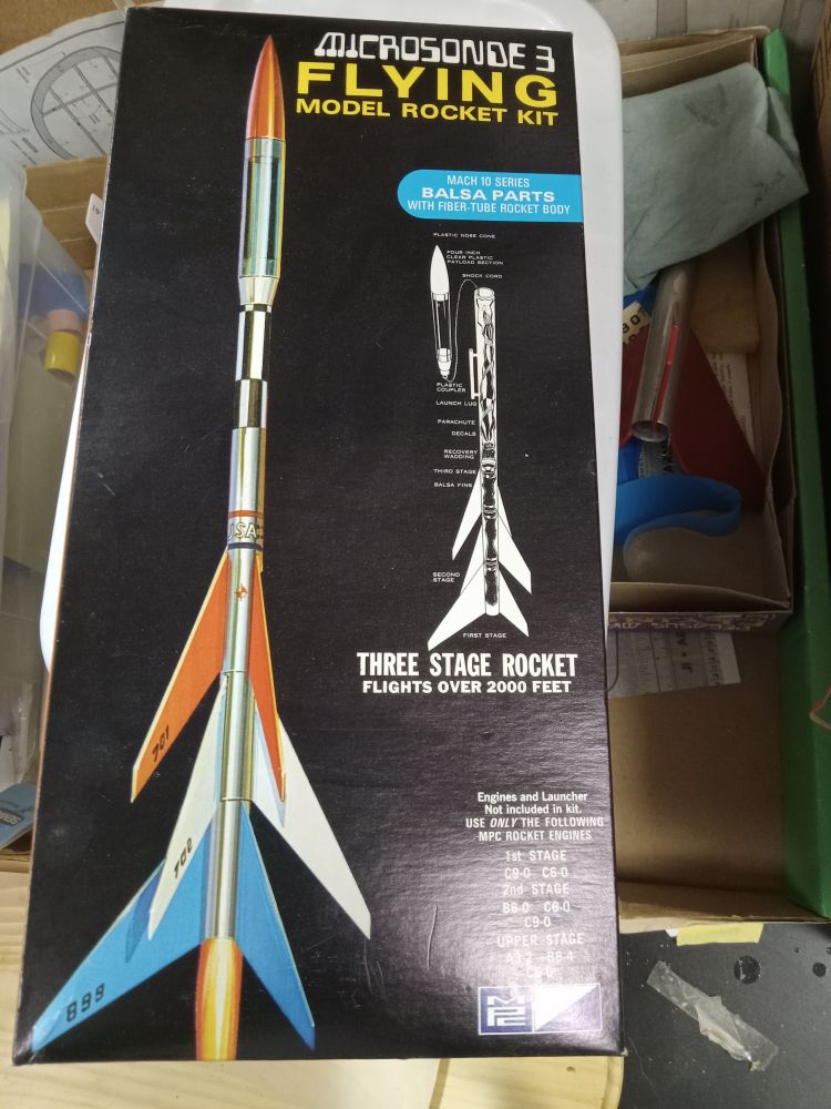 The box cover of a very old model rocket kit. It has an illustration, not a photo.  The three stage model has a silver body tube, and the 1st, 2nd, and 3rd stages have blue, white, and red fins. 