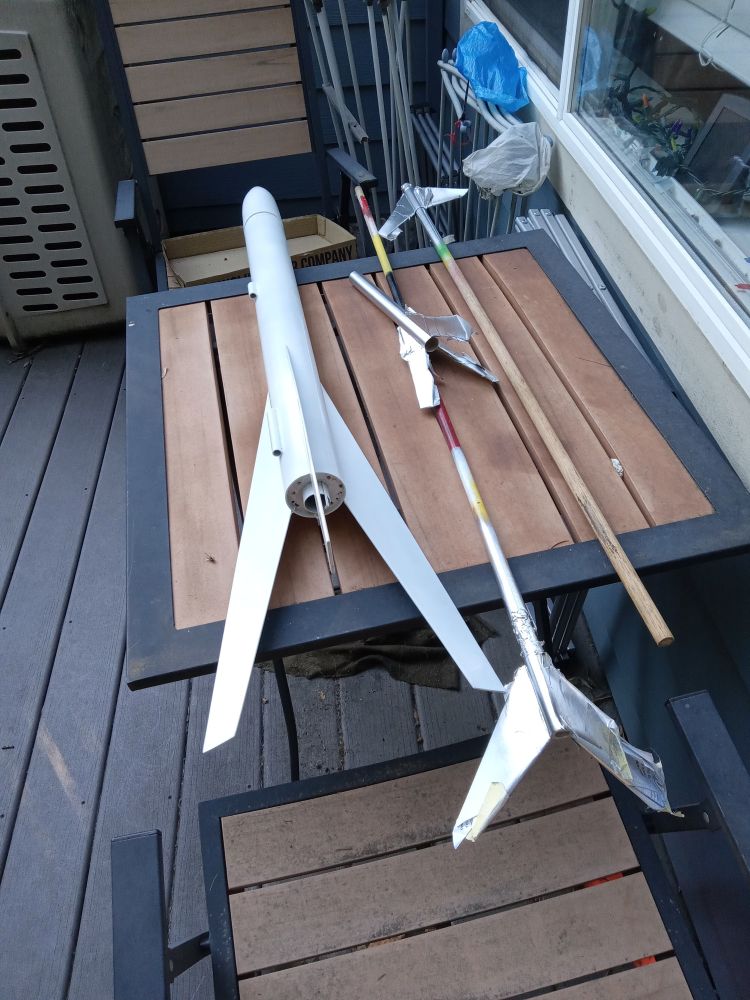 One large model rocket, and the three stages of a smaller one, freshly spray painted and left to dry in the sun on a deck.