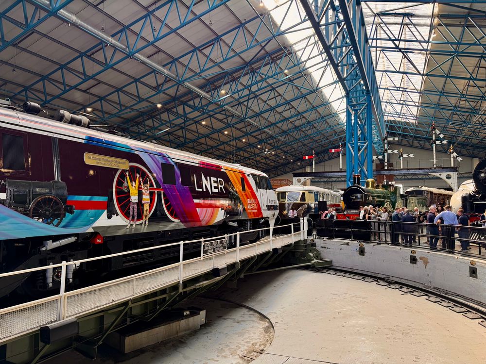 A Class 91 locomotive wearing a bright livery design of multicoloured ribbons and photographs of trains in the National Railway Museum collection, on a turntable in the main exhibition hall of the National Railway Museum in York. 