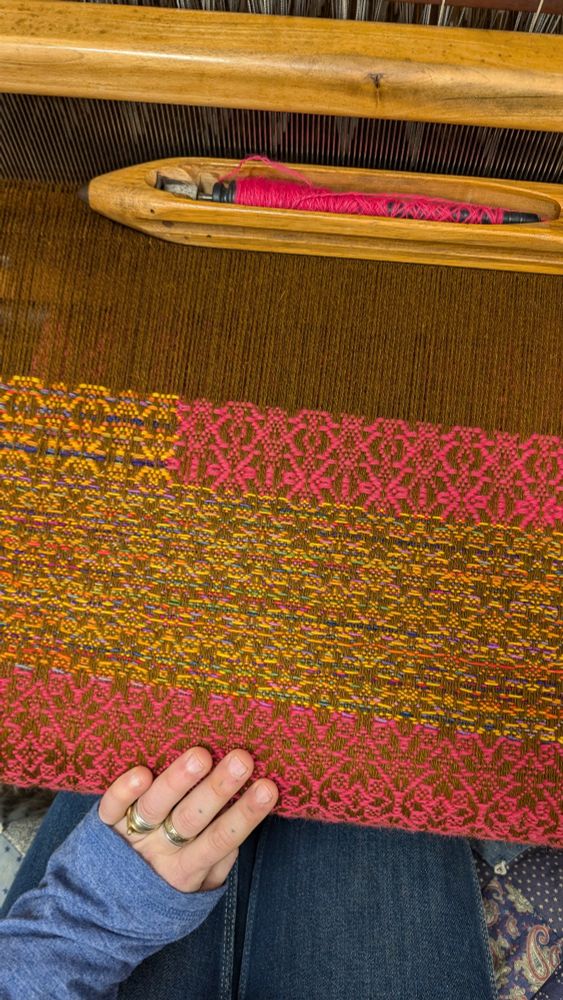 Top down view of handwoven cloth at the loom. It is wool in browns, striped yellows, and magenta with a fancy point-twill structure