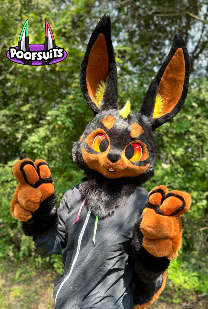 Fursuit: a friendly bunny partial with the black and rust markings of a Doberman dog, with some yellow poofs on fur on the head and inner ears. The background is a forest!