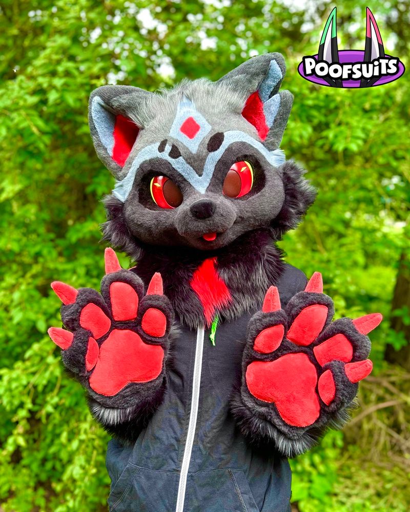 Fursuit Partial portrait: a canine creature with three tones of gray fur, accented by bright red features, like paw pads, claws, eyes, and tongue. Their paws are up, and the background is a forest.