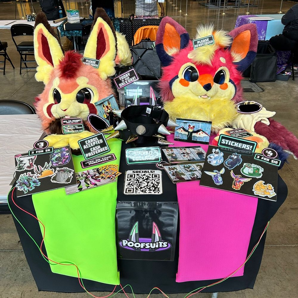 Poofsuits’ Artist Alley table at Anthrocon 2023. It has a black, pink and green theme, with plenty of stickers, portfolio photos, spiked collars, and two Fursuit partials - a bunny and a bat named Zeri and Blip