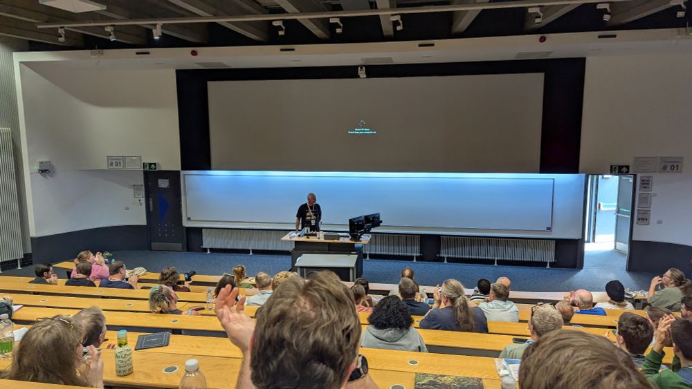 Mike Taylor standing at the front of a lecture theatre, the large screen behind him showing white text on a black backdrop reading "You're 1% there. Please keep your computer on." The people sitting in the theatre are clapping.